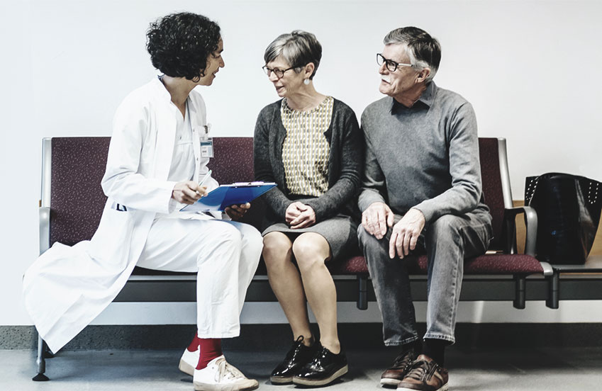 Doctor sitting on bench holding a clip board, speaking with with two patients